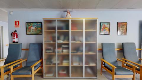 Biblioteca. Muro azul claro. El espacio está dividido en tercios con el mobiliario. Los dos tercios de los extremos tienen dos sillas largas a cada lado en azul y madera y arriba pinturas de animales. El tercio central lo ocupa un librero grande de madera con puertas de cristal opaco. Es un espacio cálido, acogedor y cómodo.