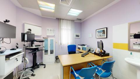 Consulta médica. Acogedora sala poligonal pintada en color lavanda. A la derecha un lavabo con espejo, seguido del escritorio. Detrás del escritorio la ventana luminosa con cortinas blancas traslúcidas y un pequeño armario blanco. Hacia la izquierda instrumentos tecnológicos de uso sanitario.