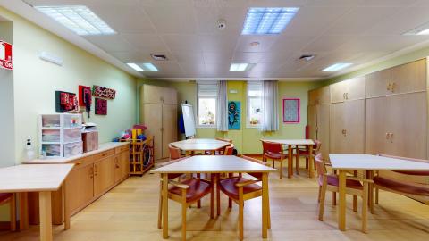 Terapia ocupacional. Sala en color verde claro con muebles en madera clara. En el muro izquierdo una credenza. En la esquina, un armario angosto. A la derecha grandes armarios de suelo a techo. Se aprecian dos ventanas luminosas con cortinas blancas y macetas. En el centro hay varias mesas con sillas que se pueden acomodar de diversas formas.
