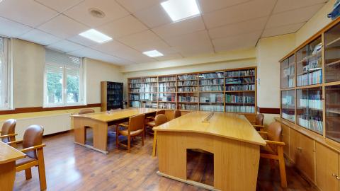 Biblioteca. Biblioteca amplia pintada en amarillo claro. En el muro de la izquierda hay dos grandes ventanas exteriores. En los muros del fondo y de la derecha hay grandes libreros de madera con puertas de madera abajo y de cristal arriba. Al centro hay mesas de lectura en madera con sillas marrones. El suelo es tarima.