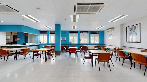 Cafetería. Amplio espacio en blanco y azul. Al fondo a la izquierda está la barra, de frente un muro con varias ventanas exteriores. Esas dos secciones están pintadas en un azul brillante, al igual que una columna intermedia. El muro de la derecha, pintado en blanco, tiene cuadros modernos en tonos azules. Al centro se distribuyen mesas y sillas en blanco y marrón.