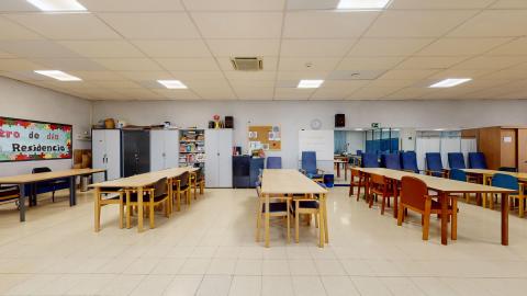 Centro de día. Amplio espacio diáfano en azules y marrones claros. En el muro del fondo hay algunos armarios azules y butacones de respaldo alto. Al centro hay cuatro mesas largas de madera con sillas para realizar las diversas actividades.