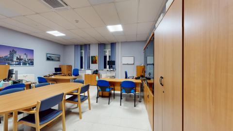 Consulta médica. Sala amplia pintada en azul con un cuadro de puente moderno en azules y morados en uno de los muros. En otro muro hay dos ventanas con cortinas azules. En el tercer muro hay armarios de madera. Se acomodan cuatro escritorios con su respectivo mobiliario y al centro una mesa para seis personas. Todos los muebles son de madera y las sillas también en tonos azules.