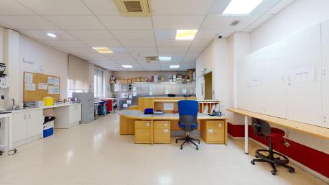 Depósito de medicamentos. Amplísima sala en blanco. Al fondo hay dos amplias ventanas exteriores. Se distribuyen varios muebles de almacenaje todo alrededor de la habitación, algunos altos y otros bajos. Al centro hay un par de escritorios.