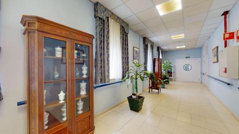 Galería. Pasillo amplio en azul claro. A lo largo hay grandes macetas, ventanas con cortinas clásicas en azules y un par de muebles vitrales clásicos de madera con jarrones cerámicos.