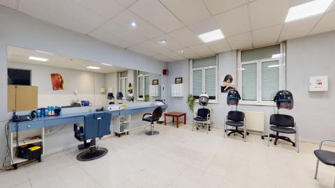 Peluquería. Amplia sala cuadrada pintada en azul claro. Del lado izquierdo hay dos estaciones de corte frente a una larga barra azul y un gran espejo corrido. Al fondo, frente a dos grandes ventanas, hay tres estaciones de secado. A la derecha, hay dos estaciones de lavado y pósters de peluquería en el muro.