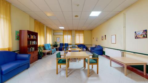 Sala de estar. Acogedora sala en amarillo claro con un librero de madera y varias zonas de estar con sofás, butacones individuales y mesas con sillas en colores azules y madera.