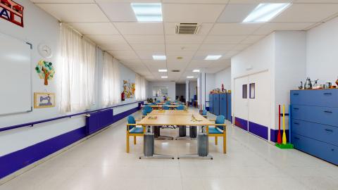 Terapia ocupacional. Sala alargada en azules claro y brillante. En el muro de la izquierda hay varias ventanas con cortinas blancas traslúcidas. En el muro de la derecha hay varios armarios de almacenaje y las puertas de acceso. Al centro hay varias mesas de madera con sillas en madera y azul que permiten varios acomodos para la realización de actividades.