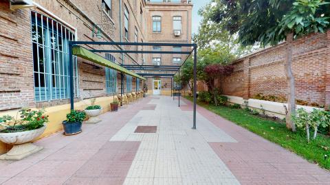 Acceso. Pasillo exterior entre la fachada delantera y la barda exterior. Toda la construcción es de ladrillo caravista. A los costados del pasillo hay jardinera y macetas. También hay un toldo plegable y sillas de exterior.
