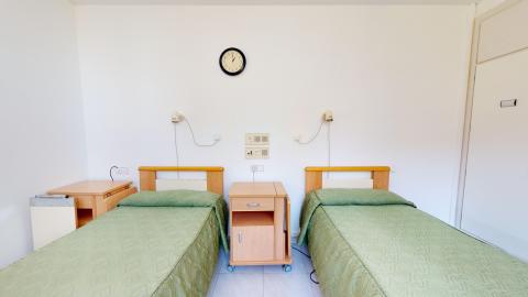 Habitación en blanco con dos camas individuales con ropa de cama en verde. Al lado de cada una hay una mesilla de noche con mesa auxiliar plegable. A la derecha, un armario empotrado con puertas blancas. 