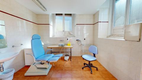 Sala en azulejos blancos con suelo anaranjado claro. Hay dos ventanas amplias y luminosas y un lavabo. La mesa de podología al centro y la silla de trabajo son ambas azules.