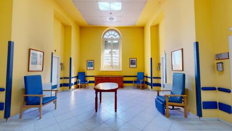 Sala de estar. Sala en amarillo con detalles en azul y butacones de respaldo alto en azul brillante. Hay una credenza en un muro y una mesa ovalada al centro, ambas de madera oscura. Los techos son muy altos y hay una gran ventana de frente.
