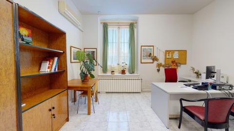 Consulta de psicología. Sala en blanco con una mesa y un librero de madera y un escritorio gris para consulta. De frente, un ventana alta muy luminosa.