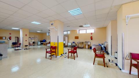Fotografía de la sala de fisioterapia. Cuenta con suelo de linóleo gris y está equipada con camillas de tratamiento, espalderas de madera en la pared, una rampa con escaleras para rehabilitación, una bicicleta estática, ruedas de hombro y balones de pilates.