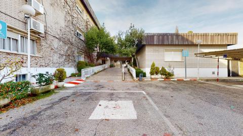 Acceso. Entrada principal exterior del centro con una rampa accesible con barandilla blanca que sube hacia la puerta. A los lados hay zonas de aparcamiento reservado con la señal de pavimento para personas con movilidad reducida y vegetación baja.