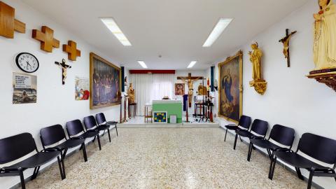 Capilla. Capilla del centro con filas de sillas negras a ambos lados de un pasillo central. Al fondo se encuentra el altar con un tapiz verde, un crucifijo y varias imágenes religiosas y cuadros colgados en las paredes blancas.