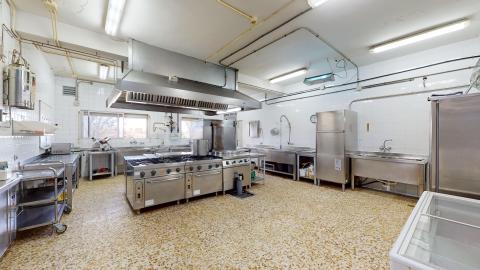 Cocina. Amplia cocina industrial con suelos de terrazo y paredes alicatadas. Cuenta con fogones de acero inoxidable bajo una gran campana extractora central, mesas de trabajo, fregaderos y electrodomésticos industriales.