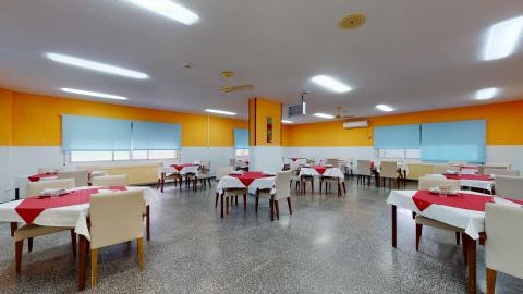 Comedor. Comedor iluminado con mesas preparadas con manteles blancos y caminos de mesa rojos. Las paredes están pintadas de color naranja y blanco, las ventanas tienen estores azules y el suelo es de terrazo oscuro.