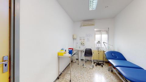 Consulta de enfermería. Consulta de enfermería con una camilla médica azul a la derecha, una mesa de acero inoxidable con material sanitario a la izquierda, un lavabo al fondo y una silla de ruedas sanitaria.