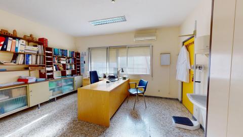 Consulta médica. Despacho médico con una gran mesa de madera, una silla de oficina azul y otra de confidente. Al fondo hay una ventana con persianas y a la izquierda una estantería llena de archivadores y libros.