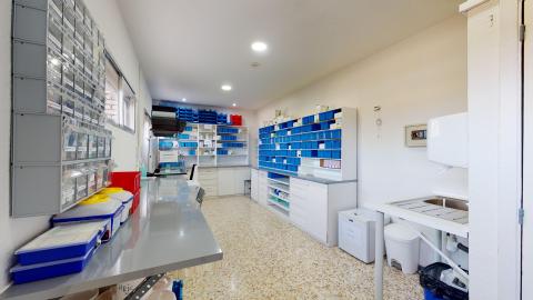 Sala de farmacia y depósito de medicamentos con estanterías y cajoneras blancas llenas de organizadores y gavetas azules. Dispone de una encimera de acero inoxidable a la izquierda y un fregadero a la derecha.