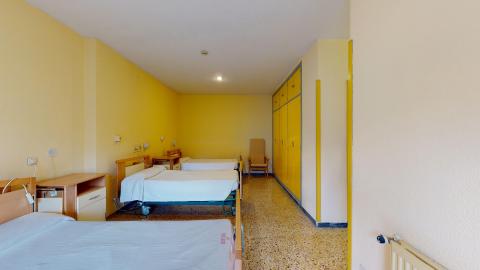Habitación triple. Habitación compartida con tres camas articuladas de hospital con sábanas blancas. Las paredes son amarillas, el suelo es de terrazo y cuenta con un gran armario empotrado emparejado con los perfiles amarillos de las paredes.
