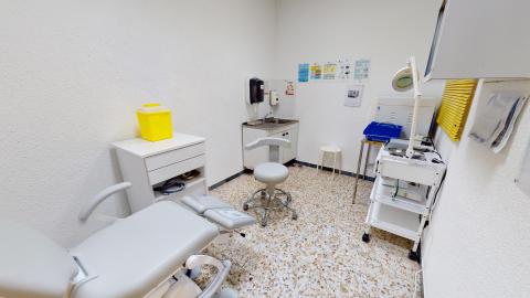 Consulta podología. Consulta de podología con un sillón clínico articulado de color azul en el centro. Cuenta con un taburete con ruedas, una lámpara de examen flexible, un lavabo y armarios con material sanitario al fondo.