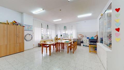 Terapia ocupacional. Sala de terapia ocupacional luminosa pintada en blanco con mesas y sillas de madera dispuestas para actividades. Las estanterías en marrón y blanco están provistas de cajas organizadoras, juegos de mesa, material didáctico y manualidades.