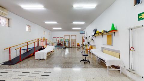 Fisioterapia. Gimnasio de fisioterapia equipado con camillas de tratamiento, paralelas de marcha, espalderas de madera en la pared, una bicicleta estática, escalerillas de dedos y diverso material de rehabilitación.