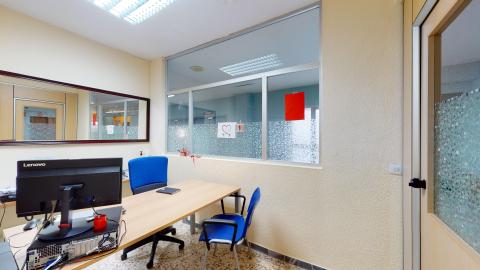 Consulta de psicología. Consulta de psicología con una mesa de oficina de madera, un par de sillas tapizadas en azul. La mitad superior de uno de los muros y la puerta de acceso son de cristal esmerilado, y detrás del escritorio hay un espejo grande a todo lo ancho, lo que da mucha luminosidad y sensación de amplitud.