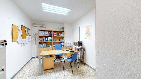 Trabajo social. Consulta de trabajo social equipada con una mesa de escritorio, ordenador, teléfono fijo y sillas para las visitas. Cuenta con archivadores de palanca en estanterías bajas y un ventanal con estor. Todos los muros son blancos.