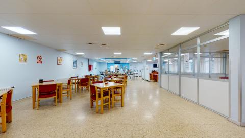 Cafetería. Zona de cafetería y descanso con mesas redondas y sillas. Cuenta con una barra de servicio al fondo, máquinas expendedoras y grandes ventanales que aportan luz natural al espacio. El muro es azul claro y el sueño de terrazo marrón claro.