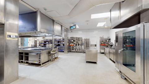 Cocina. Cocina industrial de acero inoxidable con módulos de fuegos, hornos, campana extractora de gran tamaño, mesas de preparación y fregaderos. El suelo es de baldosa industrial y las paredes están alicatadas en blanco.