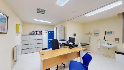 Consulta médica. Consulta médica en amarillo muy claro equipada con una mesa de escritorio principal, silla de oficina confidencial para pacientes, un módulo con cajoneras, un negatoscopio en la pared para revisar radiografías y un tallímetro con báscula. Al fondo hay archivadores y armarios.