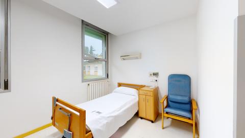 Habitación individual. Dormitorio individual con una cama articulada eléctrica con sábanas blancas y un cabecero de madera clara. Cuenta con una mesilla de noche a juego, un sillón ergonómico y un ventanal exterior que aporta luz natural. Muros, techo y suelo son blancos.