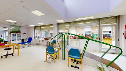Fisioterapia. Sala de rehabilitación y fisioterapia equipada con camillas regulables en altura, una rampa con escaleras y barras paralelas para el entrenamiento de la marcha, espalderas fijadas a la pared y poleas de ejercicio. Está pintado en blanco, tiene mucha luz y ventilación gracias a amplos ventanales, a un tragaluz en el techo y a que la puerta es de cristal y a al patio exterior.
