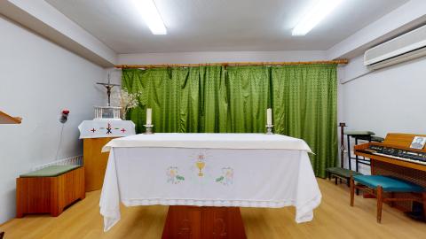Capilla. Capilla pequeña y recogida con paredes blancas y suelo de tarja de madera clara, iluminada por dos fluorescentes en el techo. En el centro destaca el altar cubierto con un mantel blanco bordado con motivos religiosos y un cáliz. Al fondo, una gran cortina de color verde oliva cubre la pared. A los lados se aprecian un sagrario con un crucifijo, un órgano electrónico de madera y un banco pequeño.