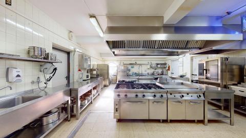 Cocina. Cocina industrial amplia y muy limpia, iluminada con luz blanca fluorescente. Predominan las superficies de acero inoxidable en encimeras, estanterías, una gran campana extractora central y los fogones de gas. Las paredes están revestidas de azulejos blancos y el suelo es de baldosas claras.