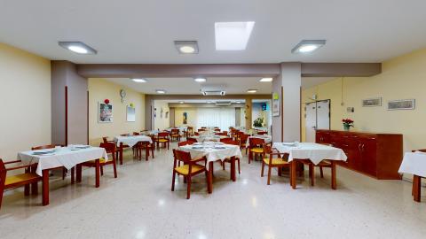 Comedor. Gran comedor rectangular con paredes de color amarillo pastel y suelo de terrazo claro pulido. El espacio cuenta con múltiples mesas cuadradas vestidas con manteles blancos y vajilla lista, rodeadas de sillas de madera con tapicería amarilla. La sala está dividida por columnas estructurales marrones y dispone de varios puntos de luz en el techo, incluyendo un tragaluz central que ilumina la zona principal. Al fondo hay una ventana amplia y luminosa.