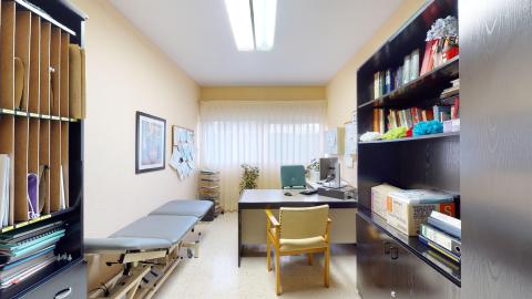 Consulta médica. Consulta médica alargada con paredes de tono salmón claro y una ventana al fondo con cortinas blancas que tamizan la luz natural. En el centro se ubica la mesa de escritorio gris con un ordenador y una silla de confidente tapizada en amarillo. A la izquierda destaca una camilla de exploración gris y un mueble archivador negro, mientras que a la derecha hay una librería oscura llena de libros y carpetas.