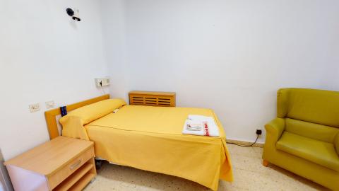 Habitación individual. Habitación individual sencilla con paredes blancas y suelo de terrazo. Cuenta con una cama articulada baja vestida con una colcha de color amarillo intenso y toallas blancas dobladas encima, donde se lee "Comunidad de Madrid". Junto a la cama hay una mesilla de noche de madera clara y, a la derecha, destaca un sillón orejero amplio tapizado en un llamativo color verde pistacho.