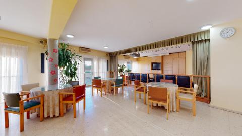 Sala de estar. Sala de estar común muy espaciosa y luminosa, con paredes de tono amarillo suave y grandes ventanales con cortinas blancas que dan a una terraza. El espacio está distribuido con varias mesas redondas vestidas con manteles de flores, rodeadas por sillones de madera con tapizados llamativos en colores rojo, verde, azul y marrón. Destaca al fondo una zona de televisión integrada en un gran mueble de madera, separada del resto de la sala por una barandilla baja y una hilera de sillones de cuero oscuro.