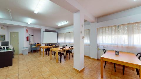 Terapia ocupacional. Aula de terapia ocupacional iluminada de forma homogénea con tubos fluorescentes en el techo. Las paredes son blancas y el suelo es de baldosas de gres en tonos arcilla. La sala cuenta con tres mesas rectangulares de madera clara alineadas, acompañadas por sillas de plástico negro. Destaca en la pared del fondo una gran pantalla enrollable blanca para proyecciones. A la izquierda se observa una mesa de despacho con ordenador y, junto a una puerta verde claro, un mueble bajo con material terapéutico.