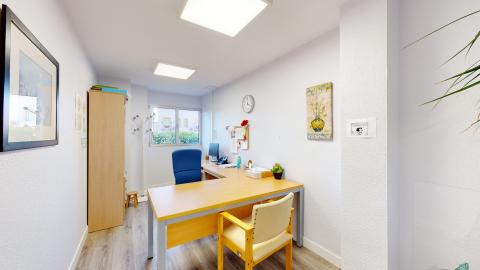 Consulta de psicología. Despacho de psicología de forma alargada, con paredes blancas y suelo de tarima gris claro. Al fondo, una ventana cuadrada aporta luz natural directa. El mobiliario principal consiste en una mesa de escritorio en forma de "L" de madera clara, con una silla de oficina ergonómica de color azul de espaldas a la ventana y una silla confidencial de madera con tapizado crema en el plano frontal. En la pared de la derecha destaca un cuadro con el dibujo de Mafalda y una pintura de un jarrón amarillo.