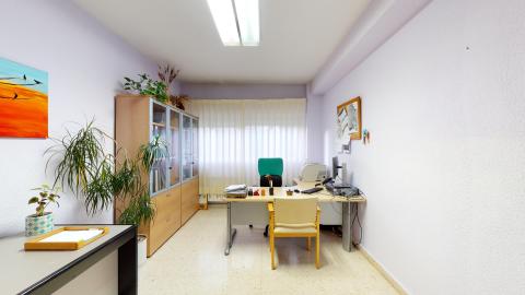 Trabajo social. Oficina de trabajo social con paredes de color lila muy suave y suelo de terrazo claro, iluminada por una ventana al fondo con cortinas de láminas verticales y un plafón fluorescente en el techo. En el centro destaca una mesa de despacho gris con una silla de oficina verde oscuro y una silla de confidente de madera clara en frente. A la izquierda resalta un gran mueble de oficina de madera con vitrinas acristaladas llenas de carpetas, decorado en su parte superior con plantas colgantes.