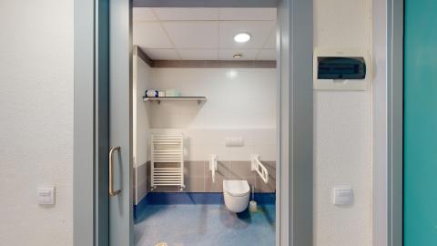 Baño y aseo. Vista frontal de un baño geriátrico adaptado a través de una puerta corredera gris. El aseo cuenta con un suelo antideslizante de color azul intenso y paredes con azulejos blancos y una franja gris a media altura. El inodoro, suspendido en la pared central, está flanqueado por dos barras de apoyo abatibles de color blanco. A la izquierda se observa un radiador toallero blanco y un estante metálico superior.