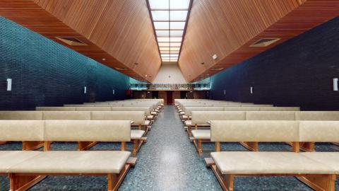 Capilla. Capilla interior de diseño moderno caracterizada por un techo monumental en forma de "V" invertida revestido de listones de madera clara, con una hilera central de tragaluces que aportan luz cenital. Las paredes laterales son de ladrillo visto de un tono gris casi negro. En el centro, un pasillo central divide dos bloques de bancos corridos de madera clara con tapicería acolchada de color beige.