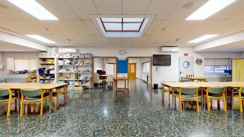 Centro de día. Sala común de actividades muy espaciosa y bien iluminada por paneles LED y un lucernario central cuadrado en el techo. Las paredes son blancas y el suelo de terrazo verde oscuro moteado. La estancia está configurada con grandes mesas rectangulares rodeadas de sillas con asientos verdes. Al fondo se ubican estanterías metálicas llenas de cajas y materiales manuales.