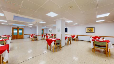 Comedor. Comedor diáfano y luminoso de paredes blancas, jalonado por columnas cuadradas y con suelo de baldosas claras. El techo técnico blanco incorpora numerosos paneles de luz cuadrados. La sala está organizada con mesas cuadradas repartidas uniformemente, vestidas con manteles blancos y cubremanteles de un llamativo color rojo, rodeadas por sillas con tapizado verde. Al fondo destaca un cuadro floral en la pared.