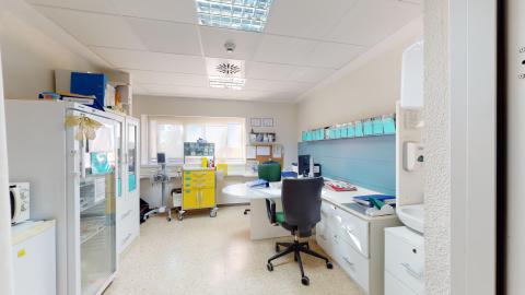 Consulta de enfermería. Espacio luminoso con suelo técnico beige y techo con paneles de luz y difusores de aire. En el centro destaca una mesa de escritorio blanca de forma semicircular con una silla de oficina negra. Al fondo, bajo una ventana con estor, hay un carro de curas amarillo brillante. El lateral izquierdo incluye una nevera de almacenamiento médico con puerta de cristal, vitrinas blancas y un lavabo con dispensador de jabón en primer plano.
